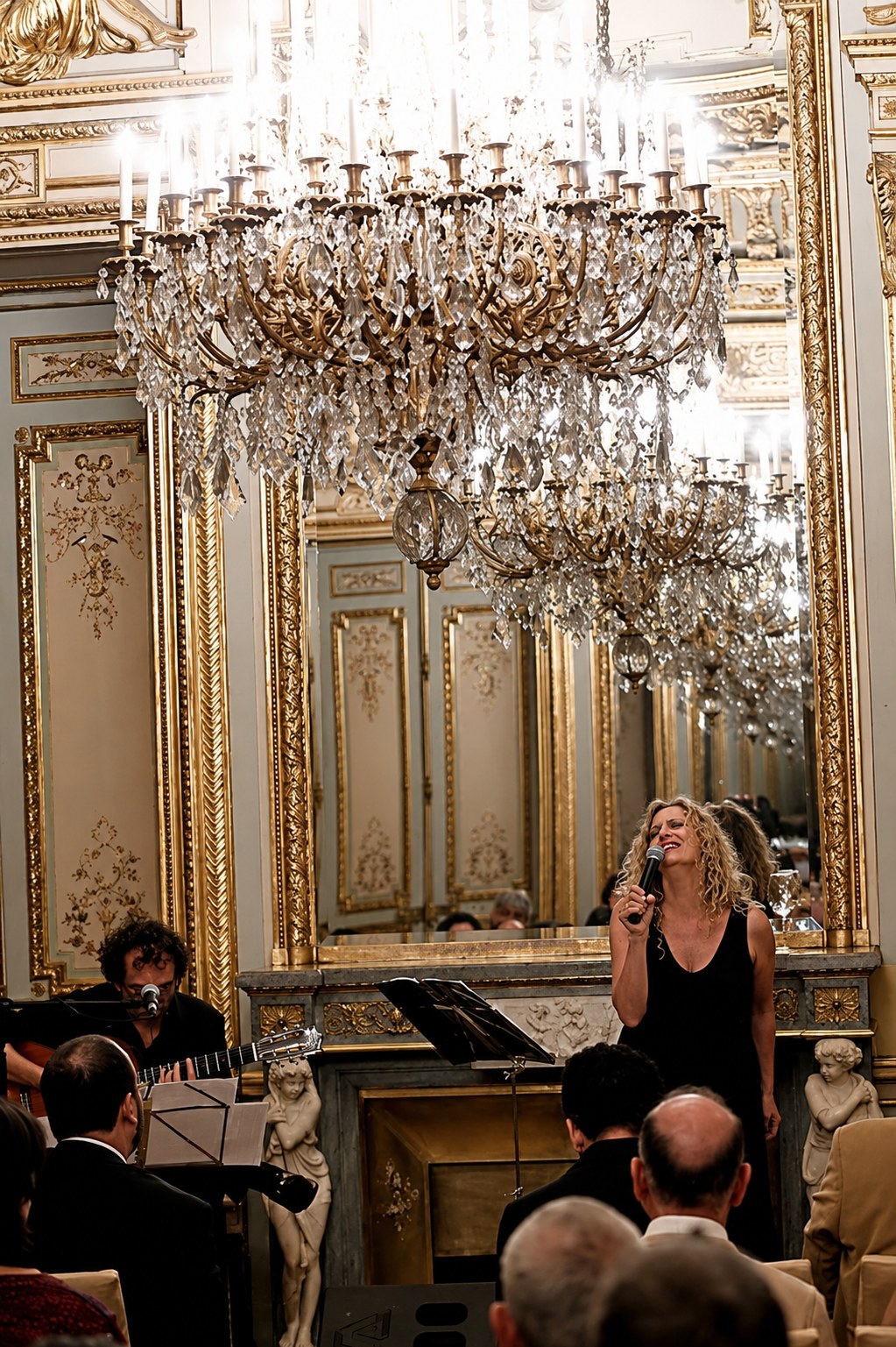 Mike y Mona en concierto: Mona cantando y Mike a la guitarra bajo una lámpara de araña en un salón dorado