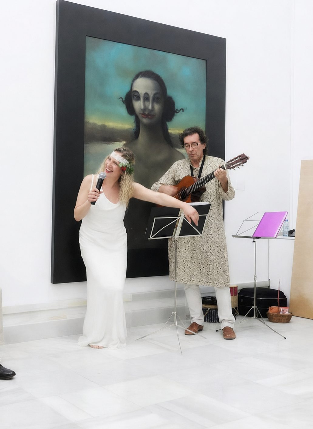 Mónica cantando con un micrófono y Miguel tocando la guitarra, delante de un gran retrato pintado.